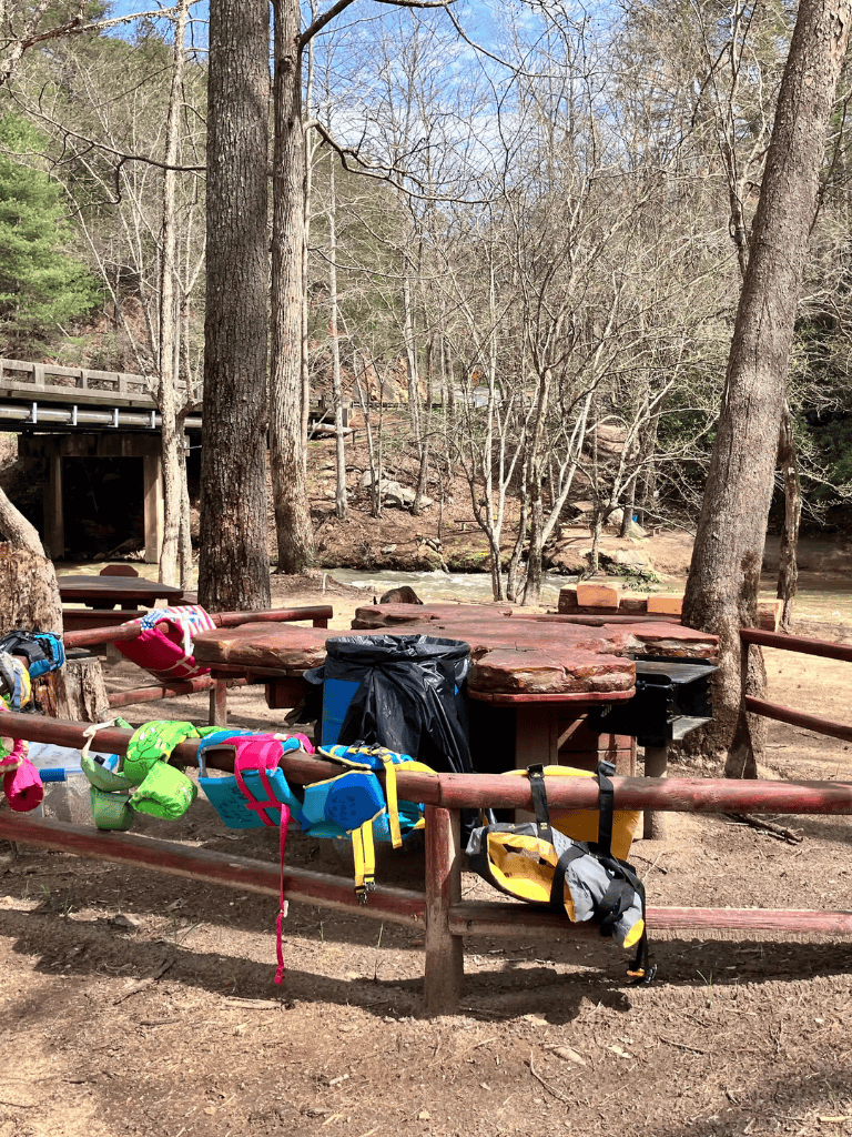 Backpacking gear and backpacks at an outdoor park picnic area with trees and a creek.