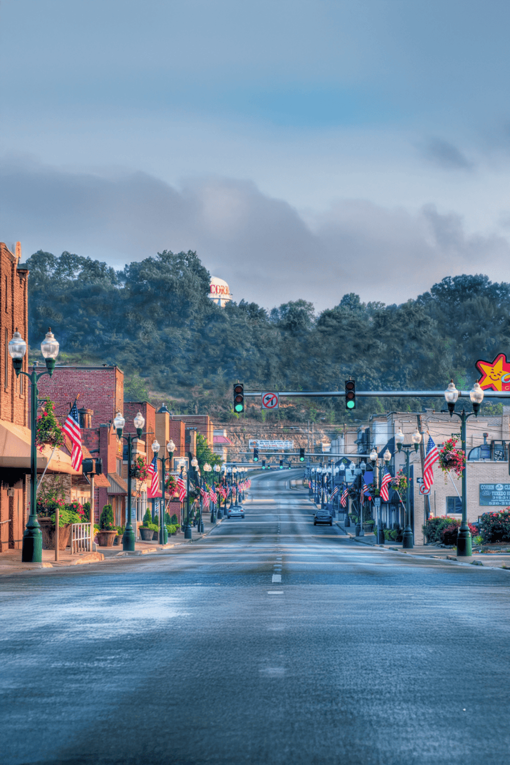 Quiet downtown street decorated with American flags and vintage lampposts, with a scenic hill and water tower in background.