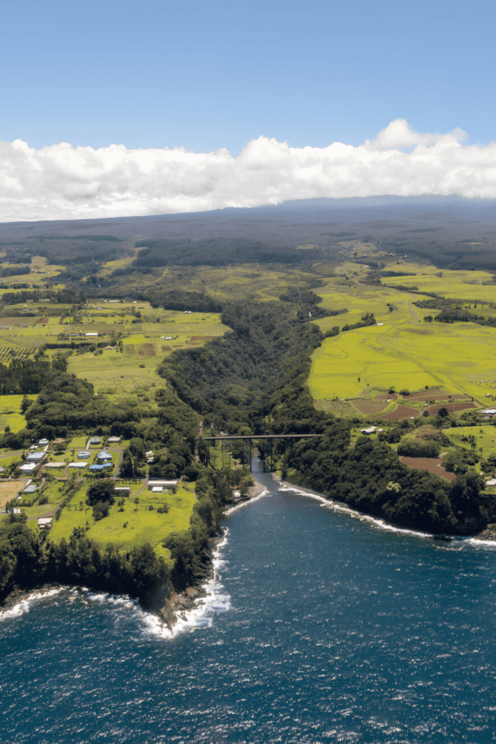 Vast green landscape with river, bridge, hills, and scattered homes under a partly cloudy sky.