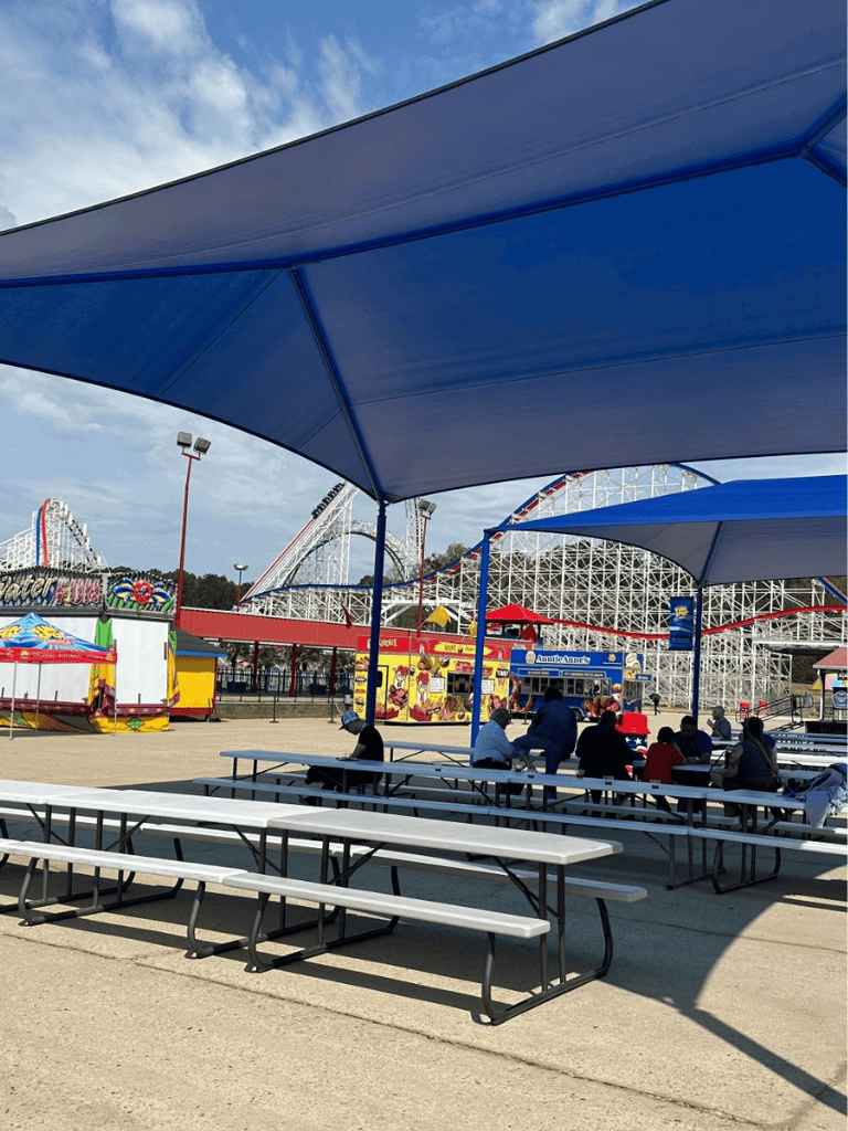 Wide view of an amusement park with roller coasters and a seating area under large blue shades.