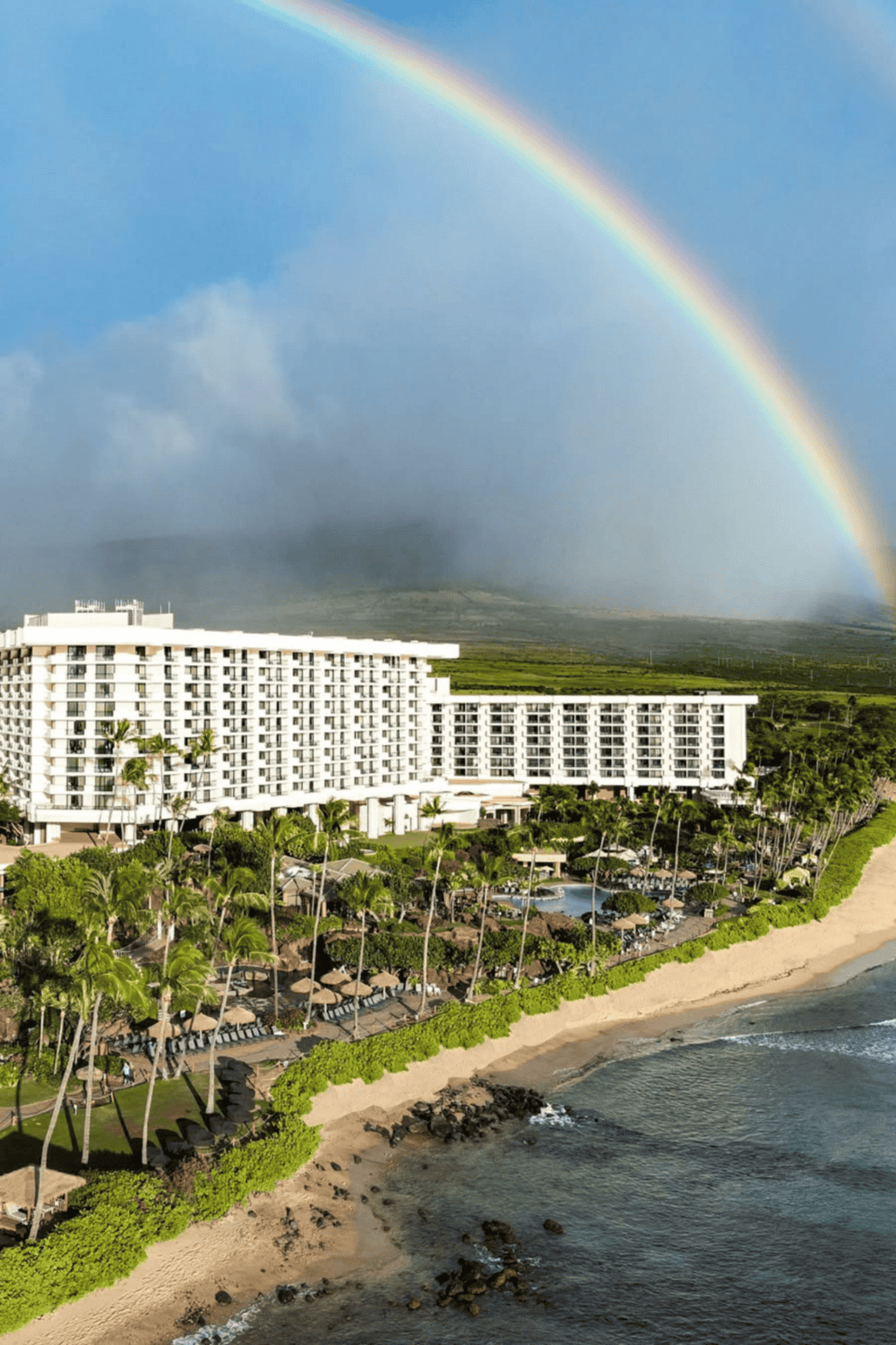 Vibrant rainbow over beachfront hotel and lush tropical landscape, Hawaii vacation destination.