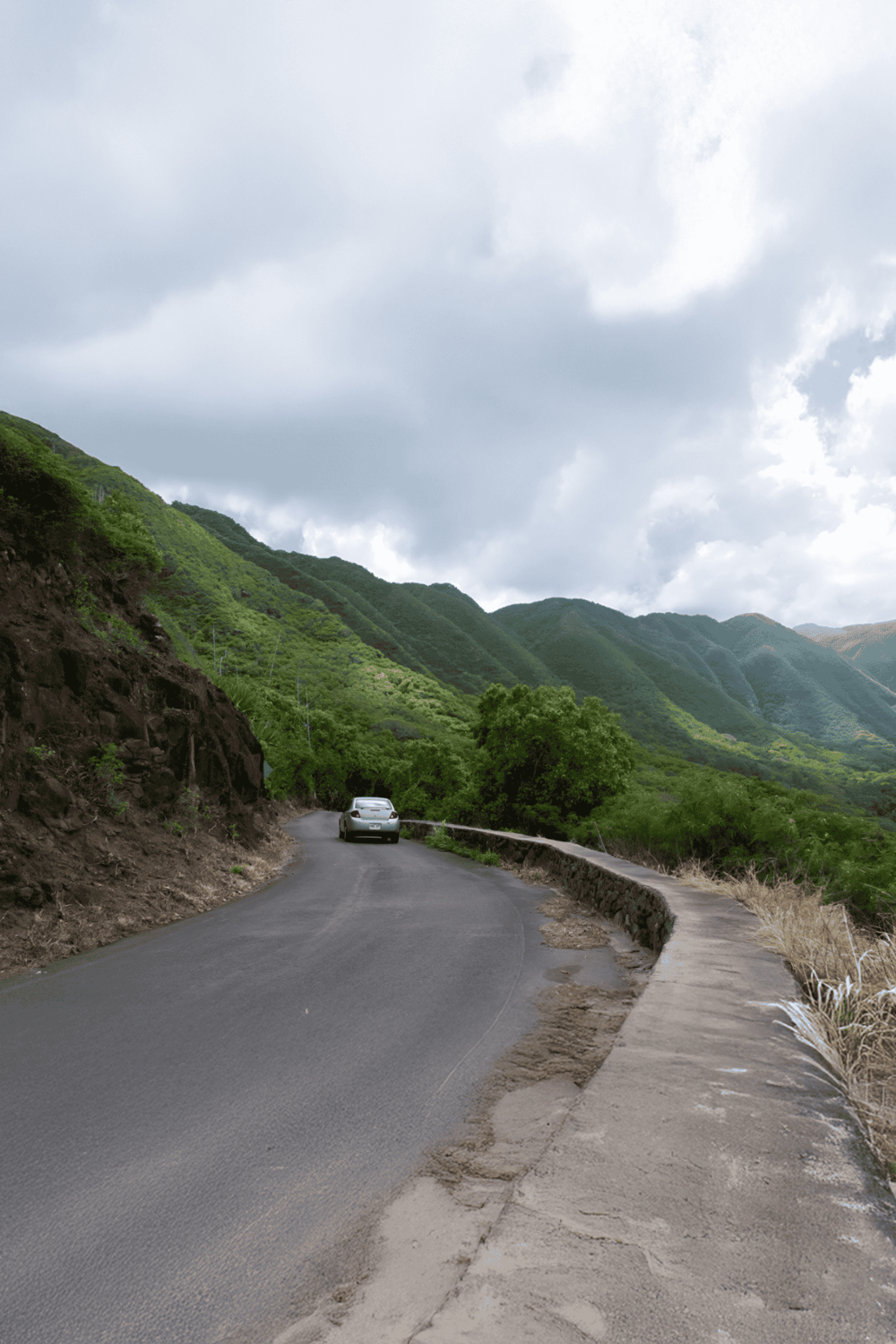 Remote mountain road with lush green hills and cloudy sky in the background.
