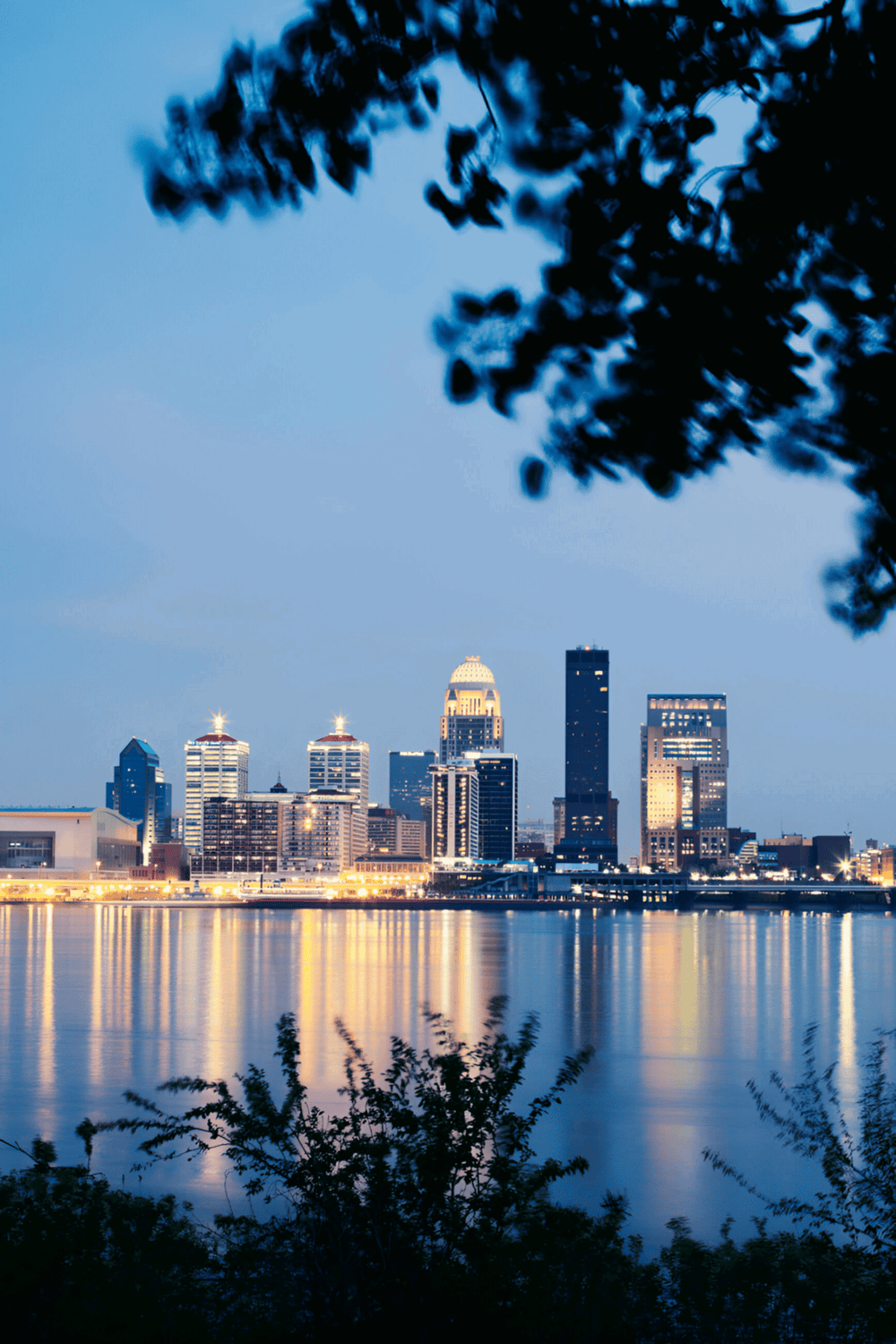 City skyline at dusk in Louisville, Kentucky, showcasing downtown skyscrapers, water reflections, and evening lighting.