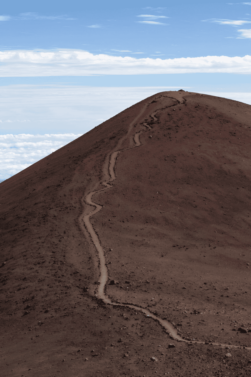 Winding dirt trail on mountain summit, offering breathtaking views of clouds and sky.