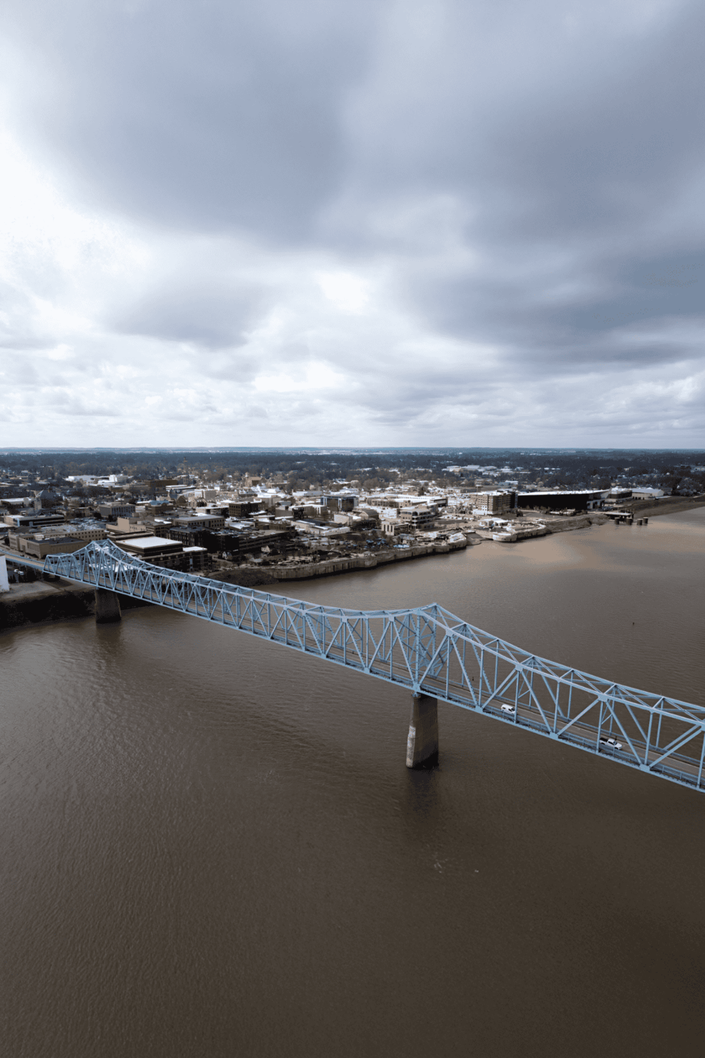 Aerial view of a city with a blue bridge crossing a river under cloudy skies, highlighting urban landscape and infrastructure.