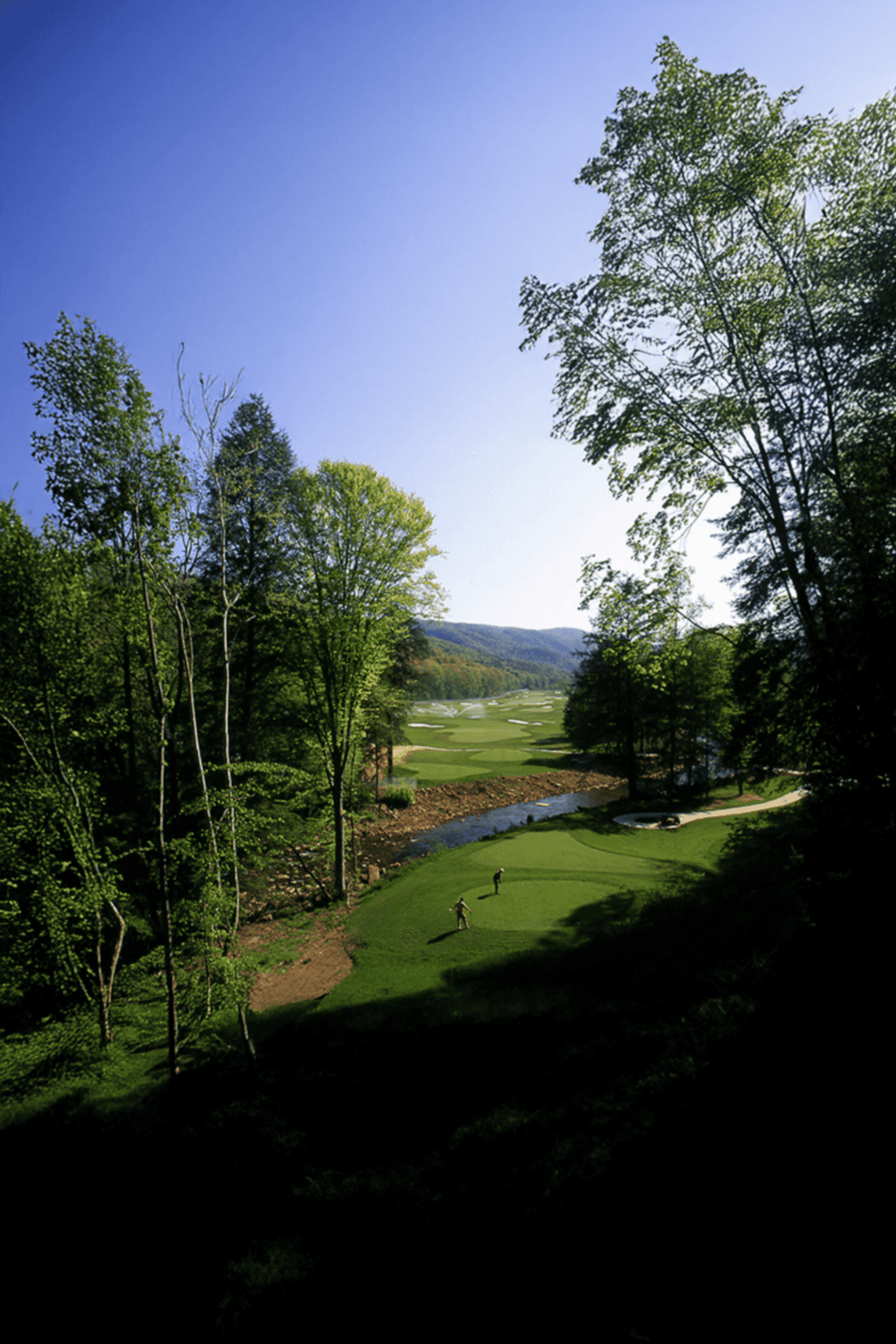 Lush green golf course surrounded by trees under a bright blue sky with rolling hills in the background.