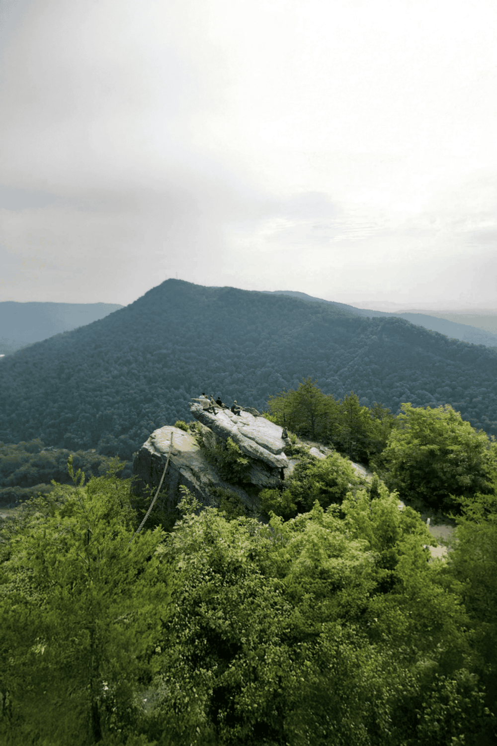 Breathtaking mountain landscape with hikers on a rocky outcrop, lush green forest, and cloudy sky.