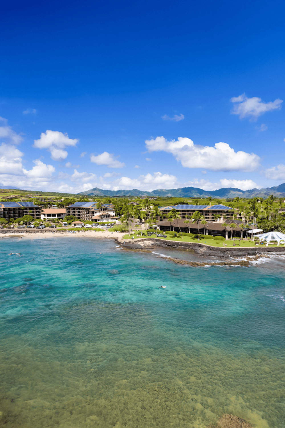 Aerial view of a beachfront resort with clear turquoise waters and lush green mountains in the background.