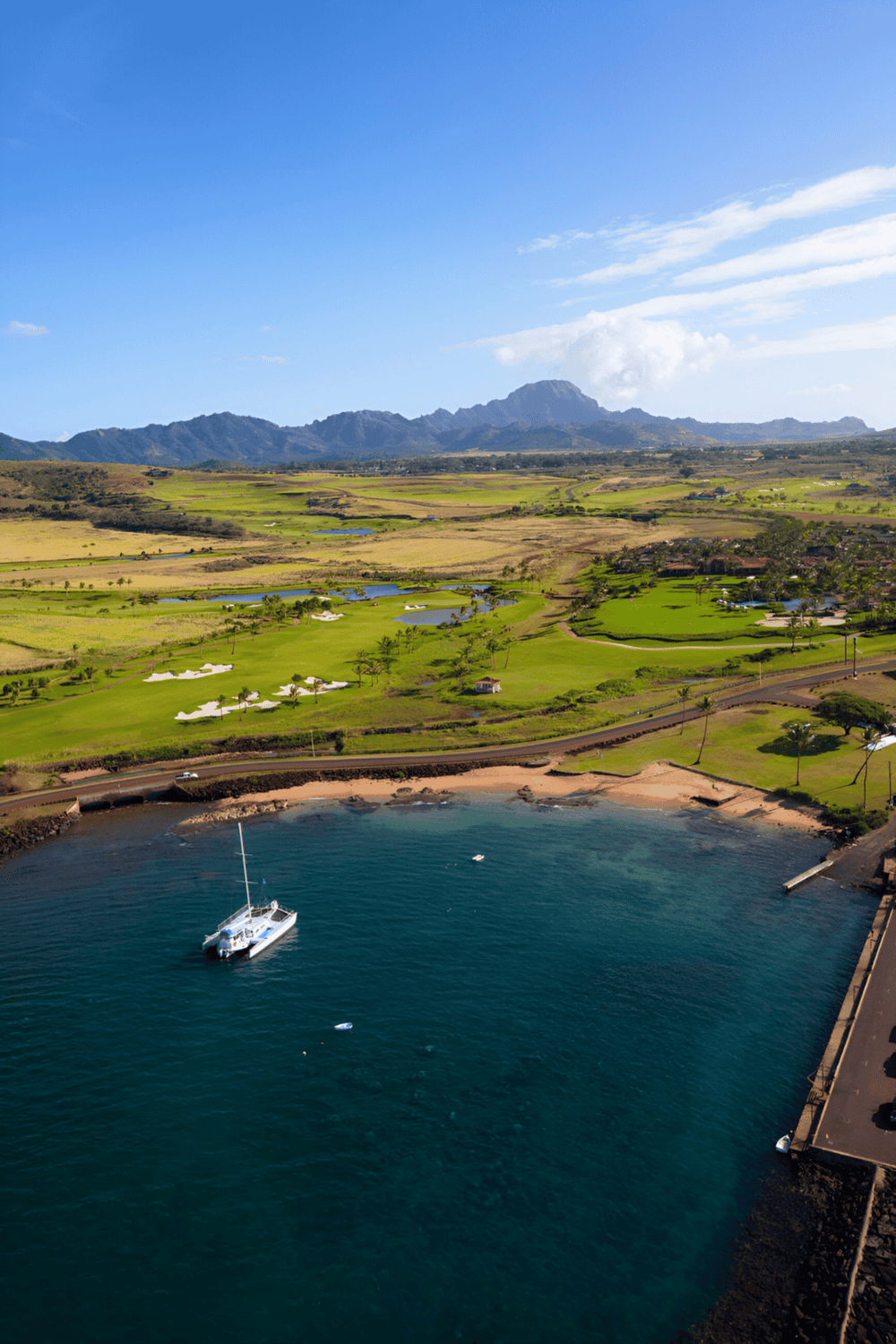 Aerial view of a scenic Hawaiian coastline with lush golf courses and mountainous backdrop.