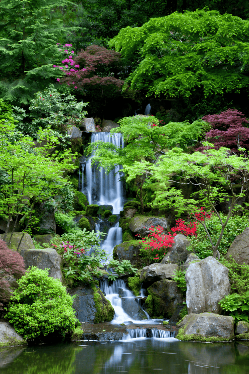 Tranquil garden waterfall surrounded by lush green trees and colorful blossoms.