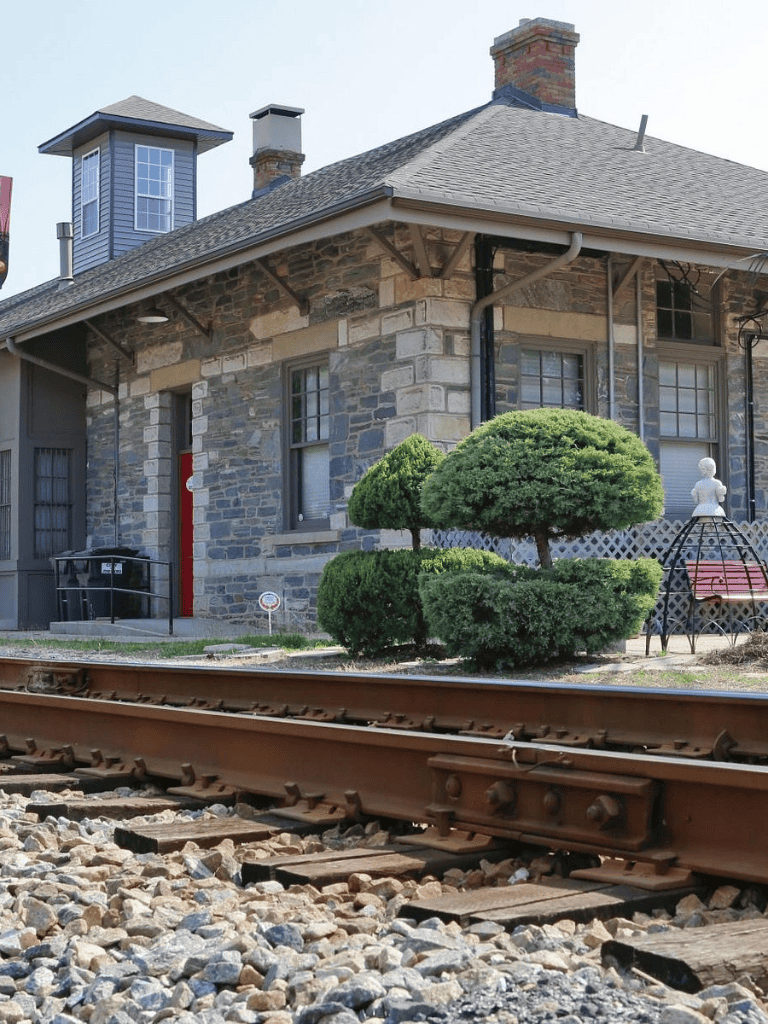 Victorian-style stone house near railroad tracks with lush shrubs and decorative garden elements.