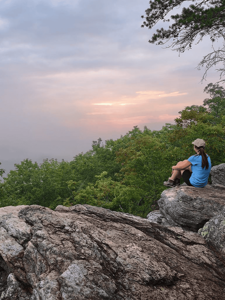 Serene woman sitting on rocky ledge enjoying sunset view over lush green trees, hiking, nature, peaceful outdoor experience.