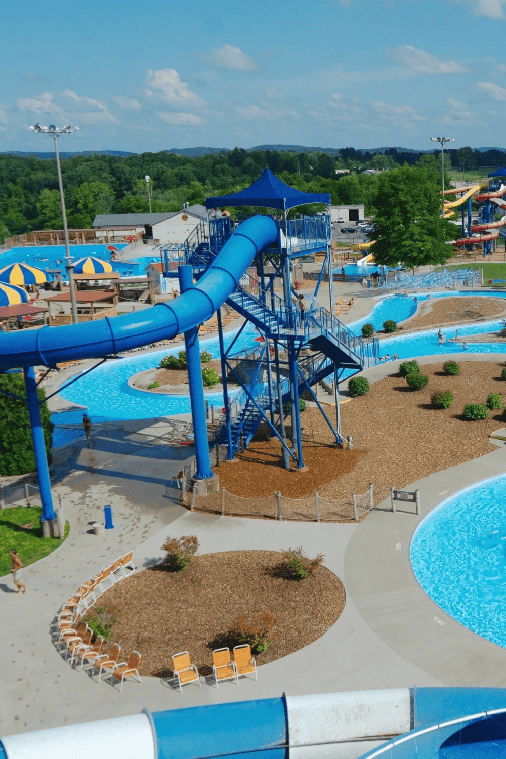 Bright blue water slides and lazy river at outdoor water park with lush greenery in the background.