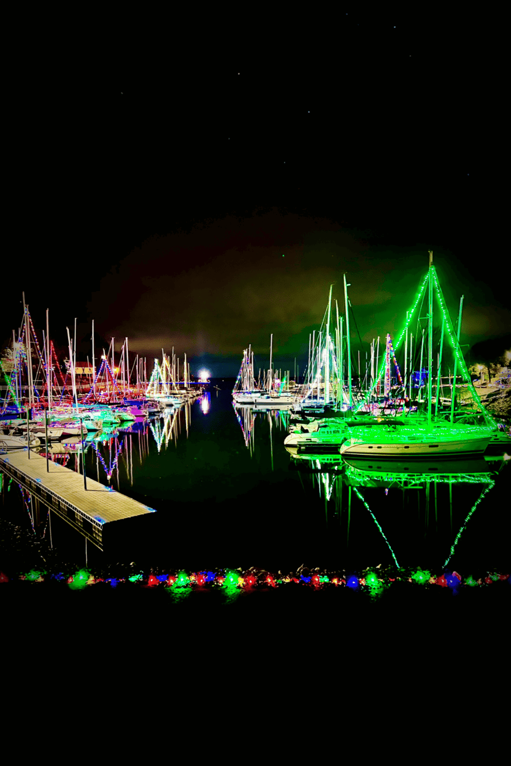 Colorful illuminated boats at night marina, festive holiday lights, waterfront scene, coastal navigation, nighttime boating illuminations.