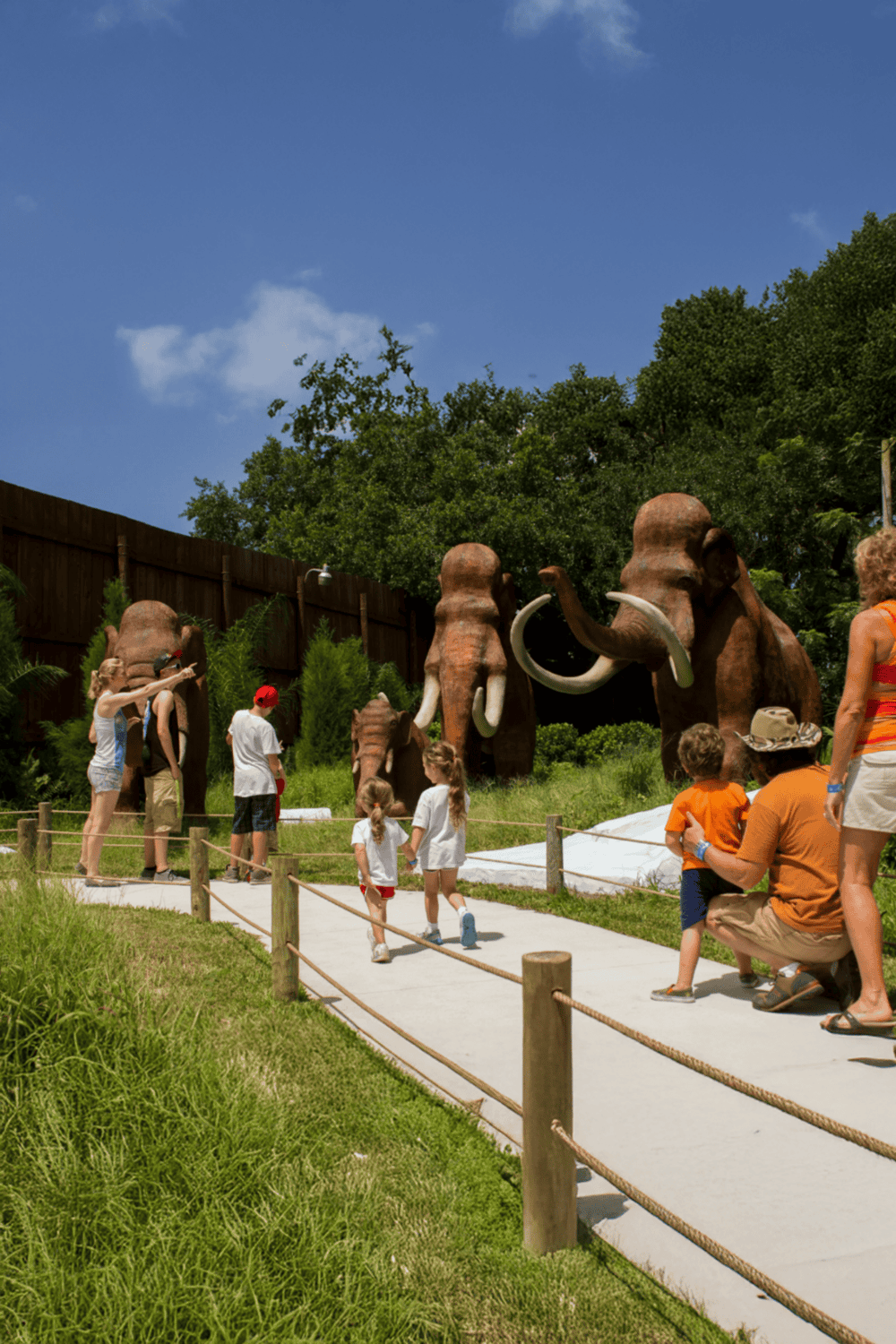 Colorful dinosaur sculptures at a zoo exhibit with children and families visiting outdoors.