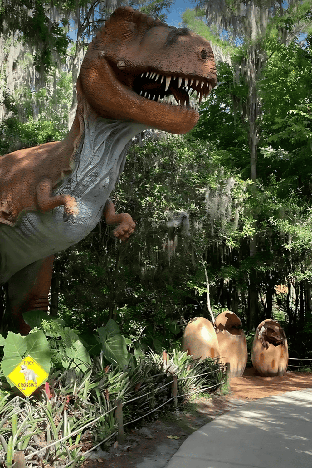 T-Rex dinosaur statue in lush outdoor park setting with egg decorations and "T-Rex Crossing" sign.