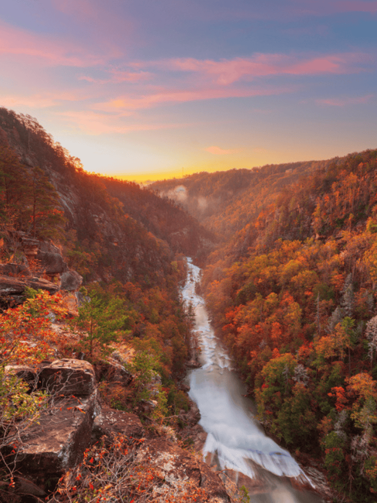 Vivid autumn landscape of a river flowing through a canyon with colorful fall foliage under a pink sunset sky.