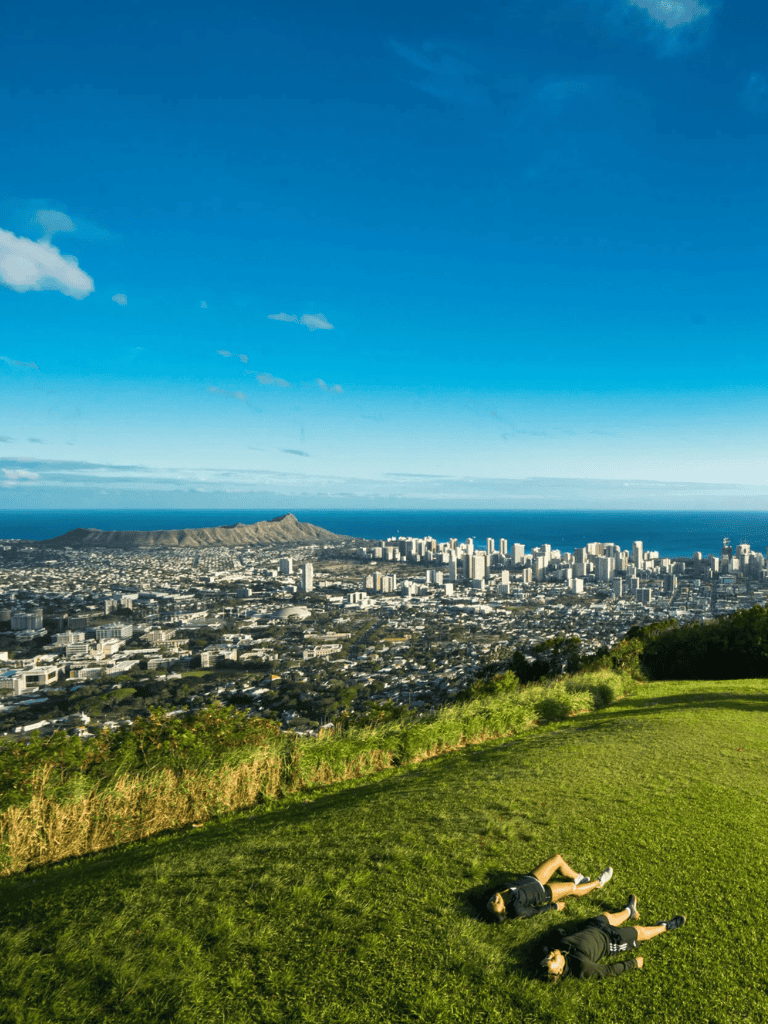 Hiking scene overlooking Honolulu skyline and mountain under blue sky, lush green grass, adventure travel.