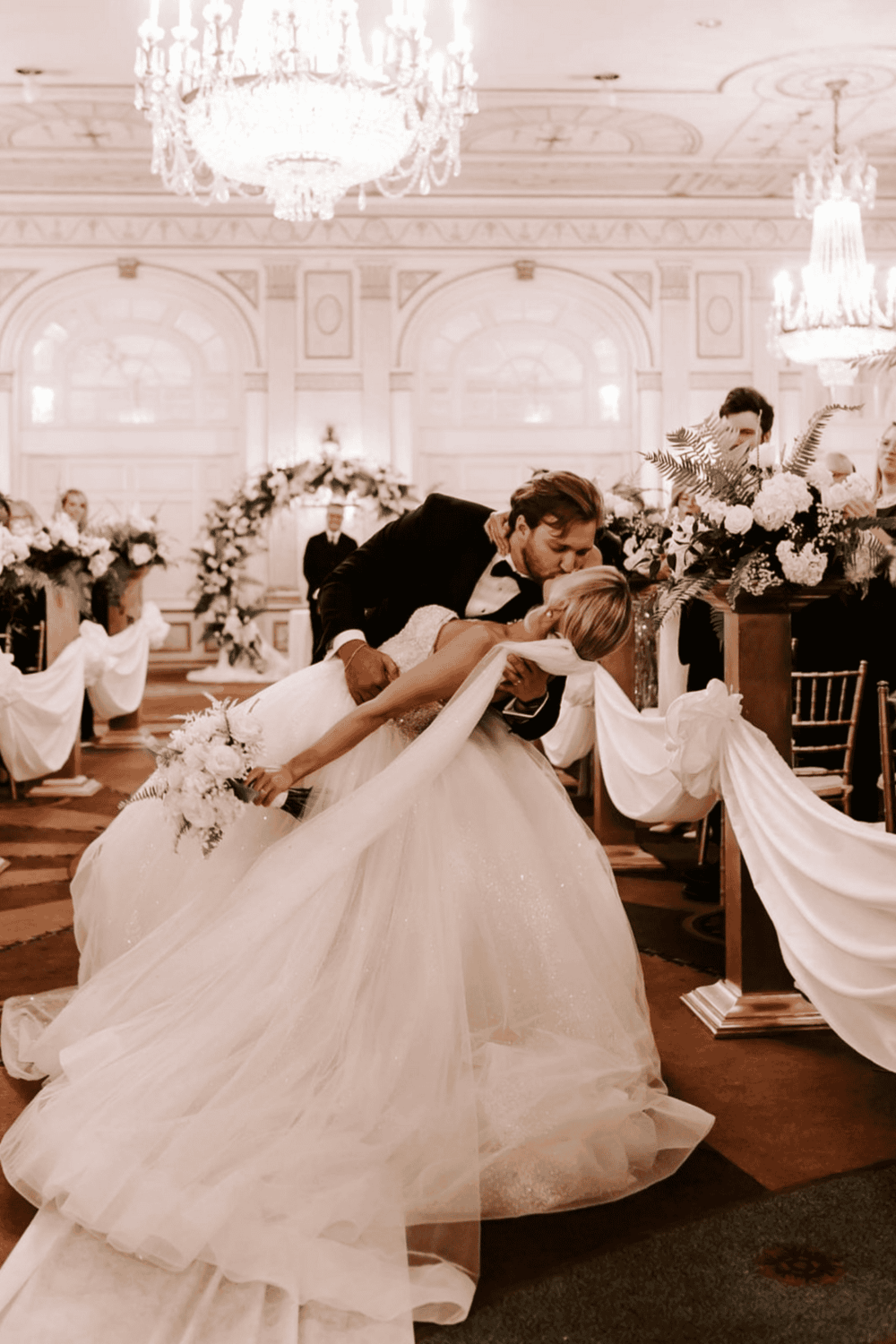Elegant wedding scene in luxurious ballroom with bride and groom, wedding vows, floral decor, and chandeliers.