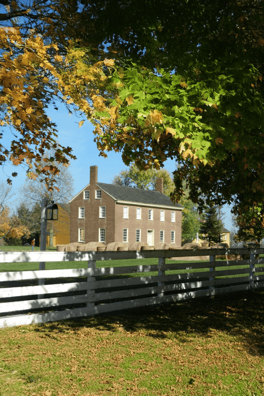 Vintage brick house under autumn trees with green leaves and white fence, scenic neighborhood view.