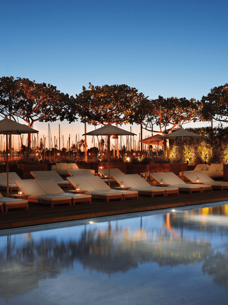 Sunny outdoor poolside lounge area at sunset with palm trees and boats in the background, perfect for relaxation and travel.