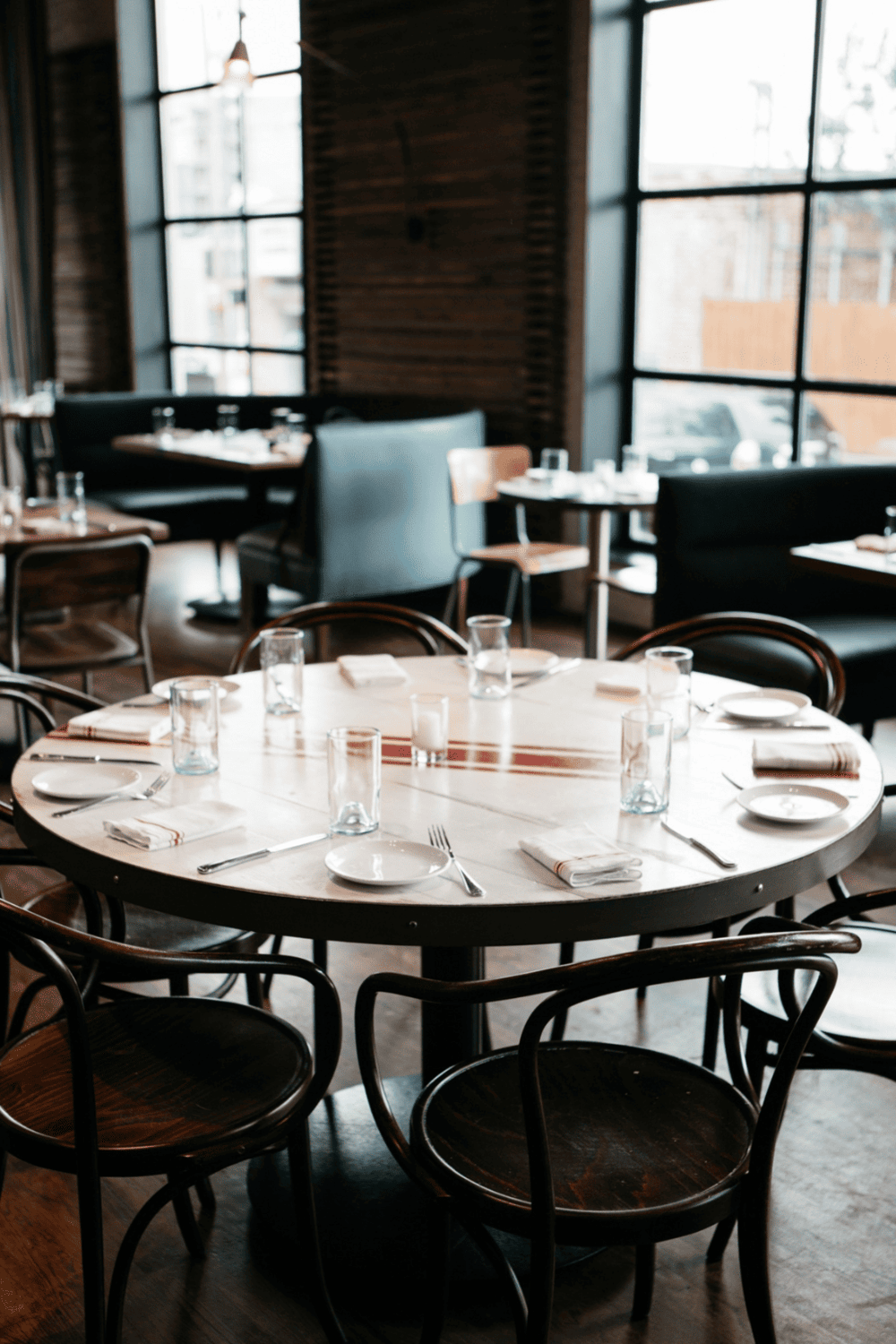 Cozy restaurant dining area with round table, neatly set with cutlery and glasses, modern interior design with large windows.