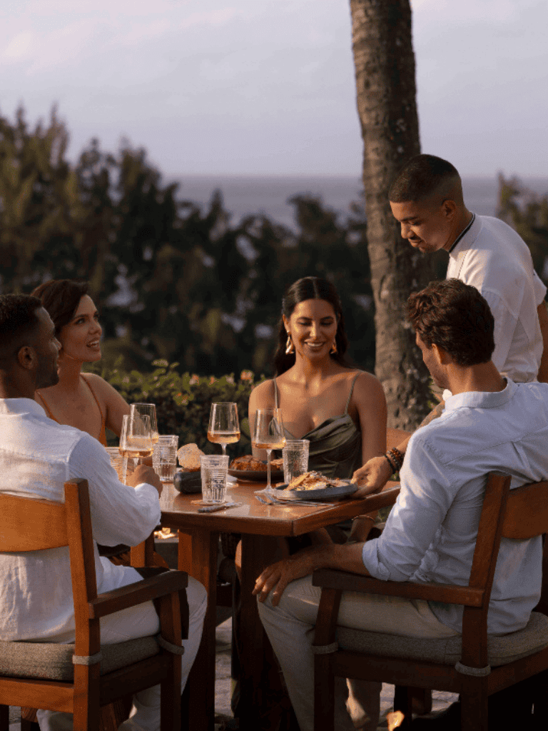 Relaxed outdoor dinner with friends at sunset, tropical scenery, wine glasses, elegant evening dining experience.