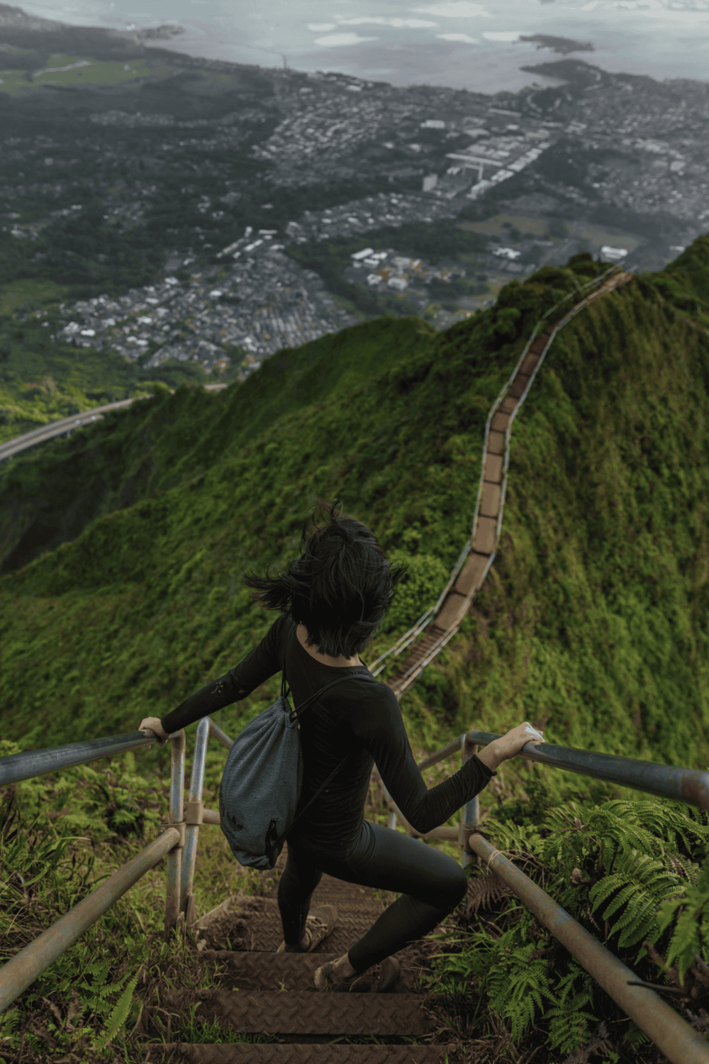 Climber ascending a lush green mountain trail with panoramic city view in the background.