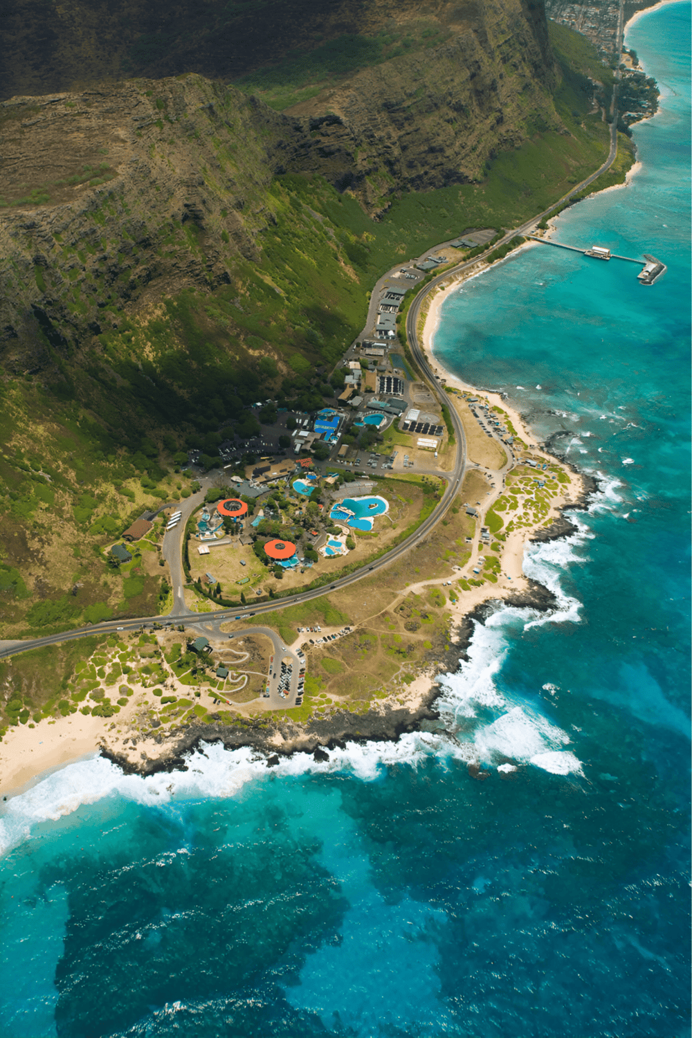 Aerial view of a coastal resort with pools, situated along a rugged shoreline and green cliffs.