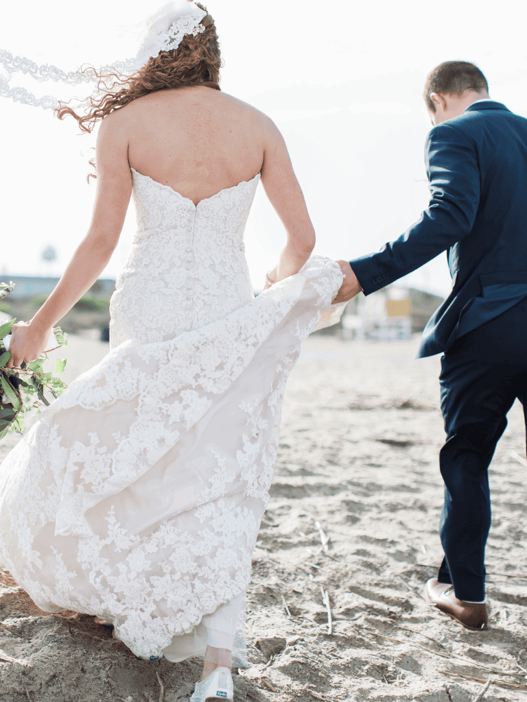 Elegant bride in lace wedding dress and groom in suit walking on beach for wedding photo.