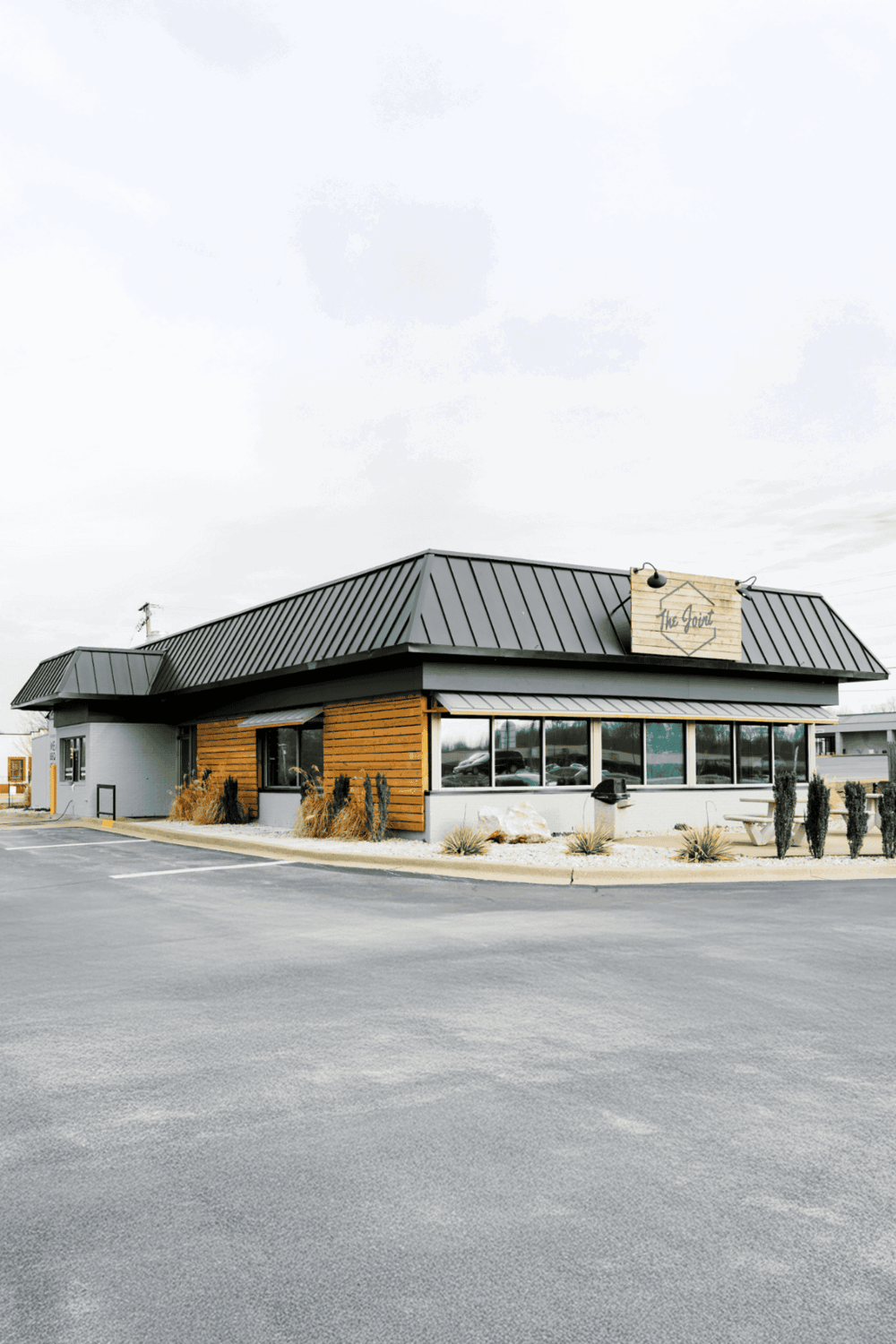 Modern restaurant exterior with wood and metal accents, featuring large windows and outdoor seating, in a commercial area.