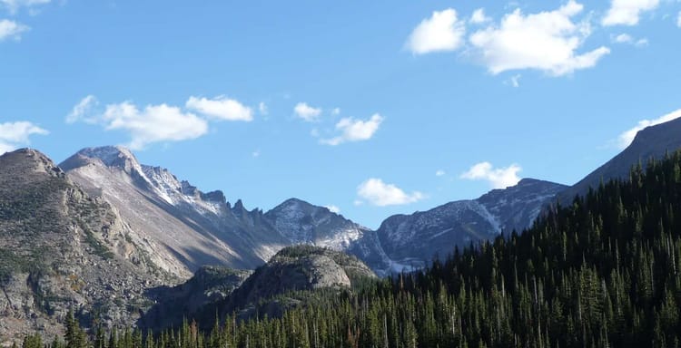 Serene mountain lake with forested shoreline and snow-capped peaks under blue sky.