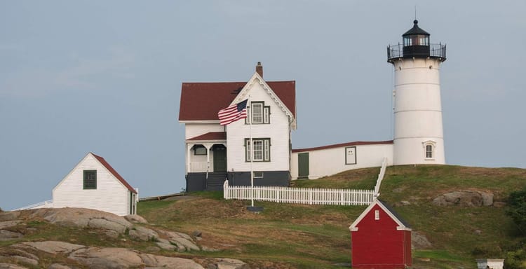 Sunny lighthouse with a historic house and outbuildings on rocky coast, scenic maritime landmark.