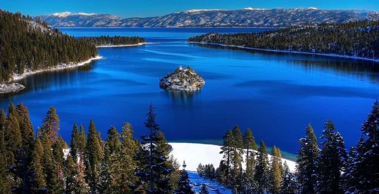 Snow-covered mountains reflecting on a calm blue lake surrounded by evergreen forests in winter.