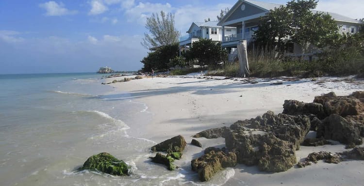 Tranquil beachfront homes facing turquoise waves and white sand under a bright partly cloudy sky.