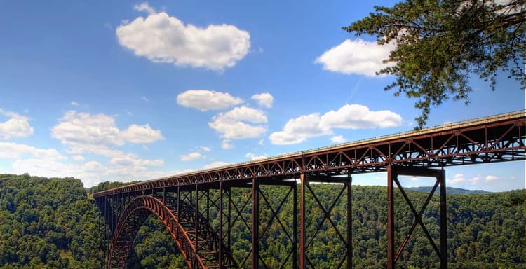 Rustic iron bridge over lush green forest with blue sky and clouds, scenic transportation route.