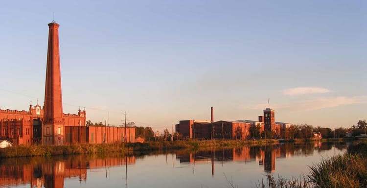Vintage industrial buildings along a calm river at sunset, reflecting a historic factory setting.