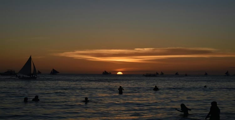 Sunset over the ocean with sailing boats and people swimming at dusk.