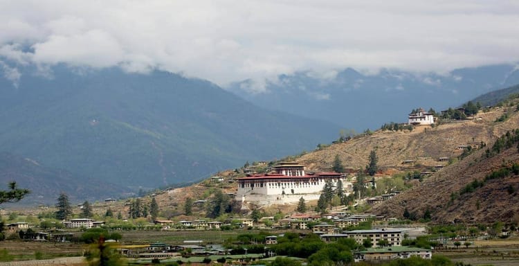 Traditional Himalayan monastery nestled on a mountain hillside in Bhutan, surrounded by green and brown terraced fields.