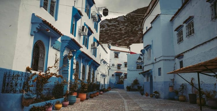 Colorful blue and white Mediterranean street with potted plants and mountain backdrop.