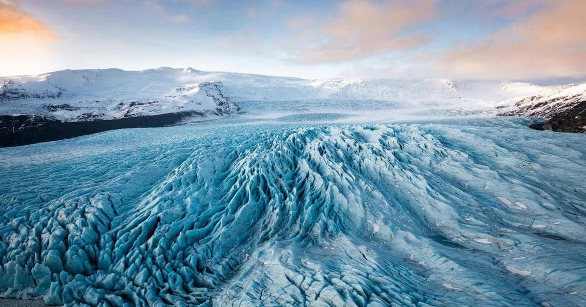 Mighty glacier in Iceland with snow-covered mountains and vibrant sky in the background.