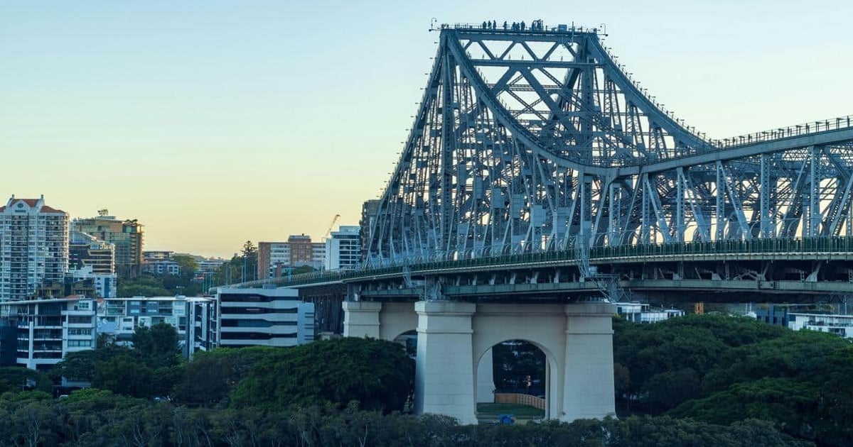 Steel truss bridge in Brisbane, Australia, over lush green trees with city skyline background.