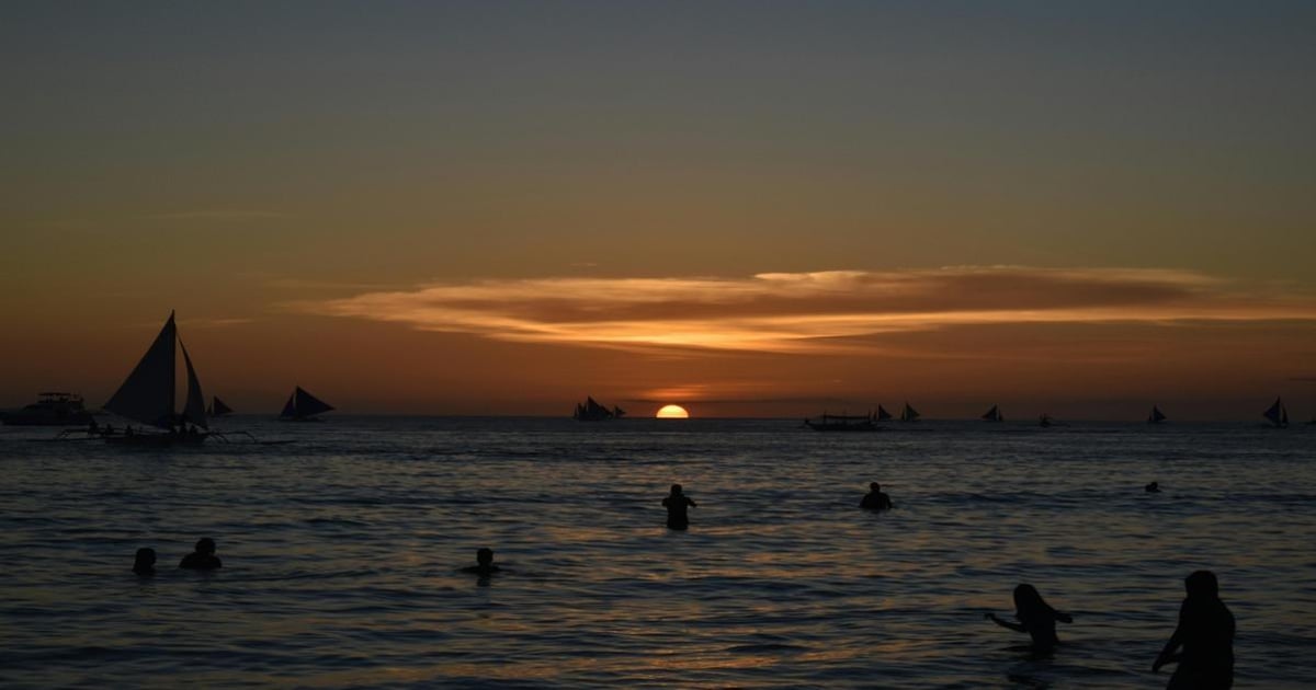 Sunset over the ocean with sailing boats and people swimming at dusk.