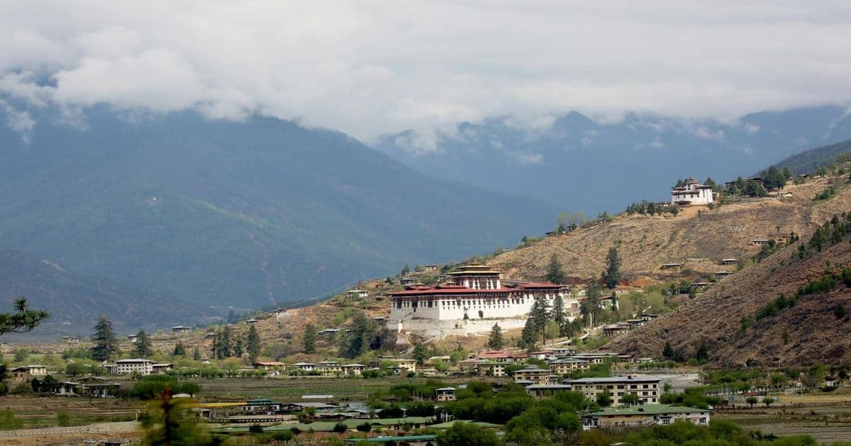 Traditional Himalayan monastery nestled on a mountain hillside in Bhutan, surrounded by green and brown terraced fields.