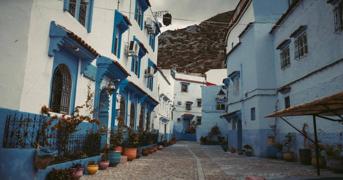 Colorful blue and white Mediterranean street with potted plants and mountain backdrop.